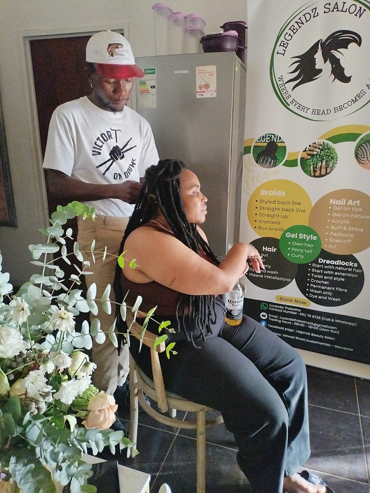 Stylist working on a client's long locs inside Legendz, beside the salon's service banner